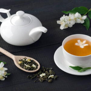 Cup of tea with jasmine flowers and teapot on dark wooden background.