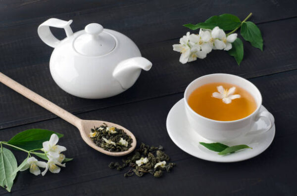Cup of tea with jasmine flowers and teapot on dark wooden background.